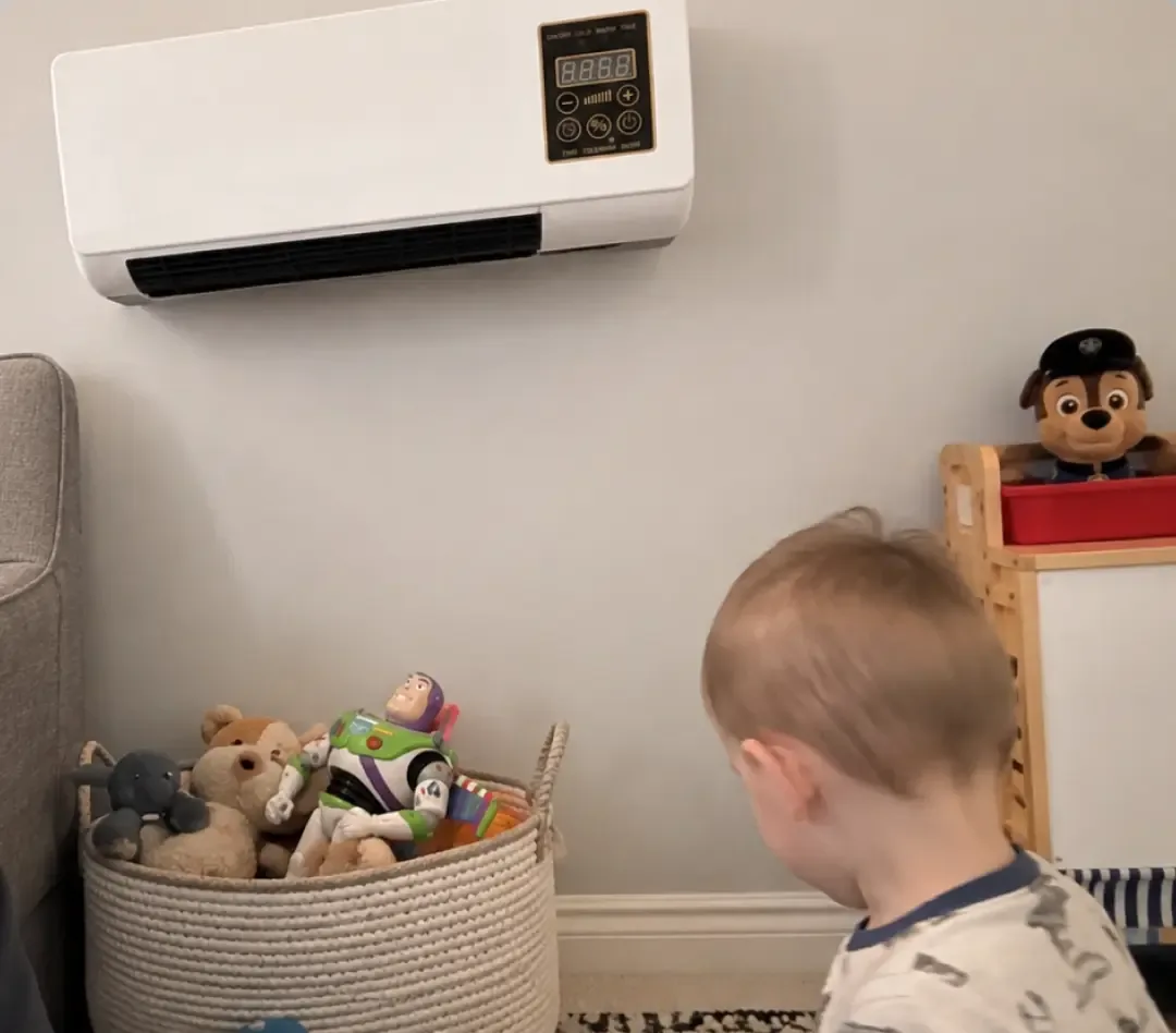 A toddler playing on the floor below the white heater mounted high on the wall